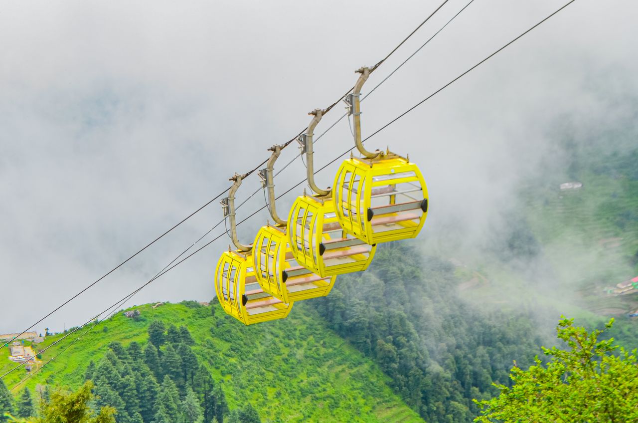 gallery-2 Bright yellow cable cars glide over lush green mountains in a misty, serene setting.